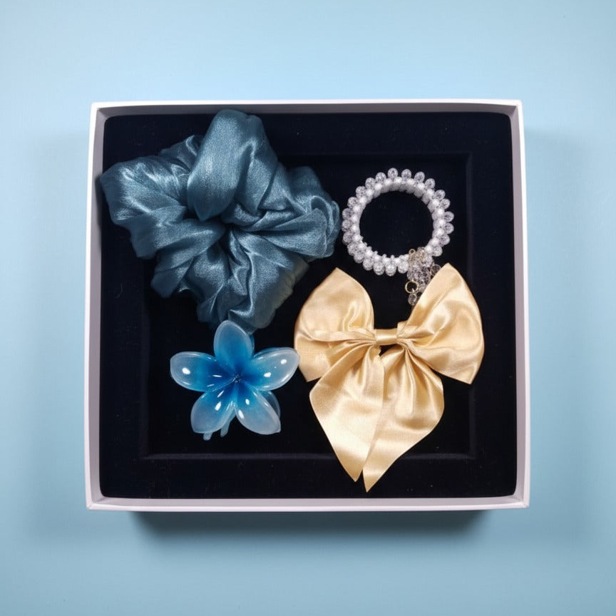 Set of hair accessories including a blue scrunchie, gold bow, and blue flower in a box on a light blue background.